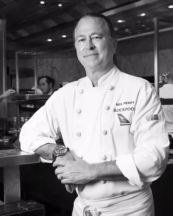 Black and White portrait of Neil Perry in a commercial kitchen leaning on the bench looking at the camera.