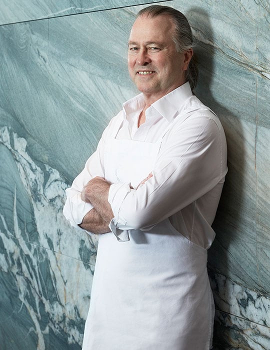 Black and White portrait of Neil Perry in a commercial kitchen leaning on the bench looking at the camera.